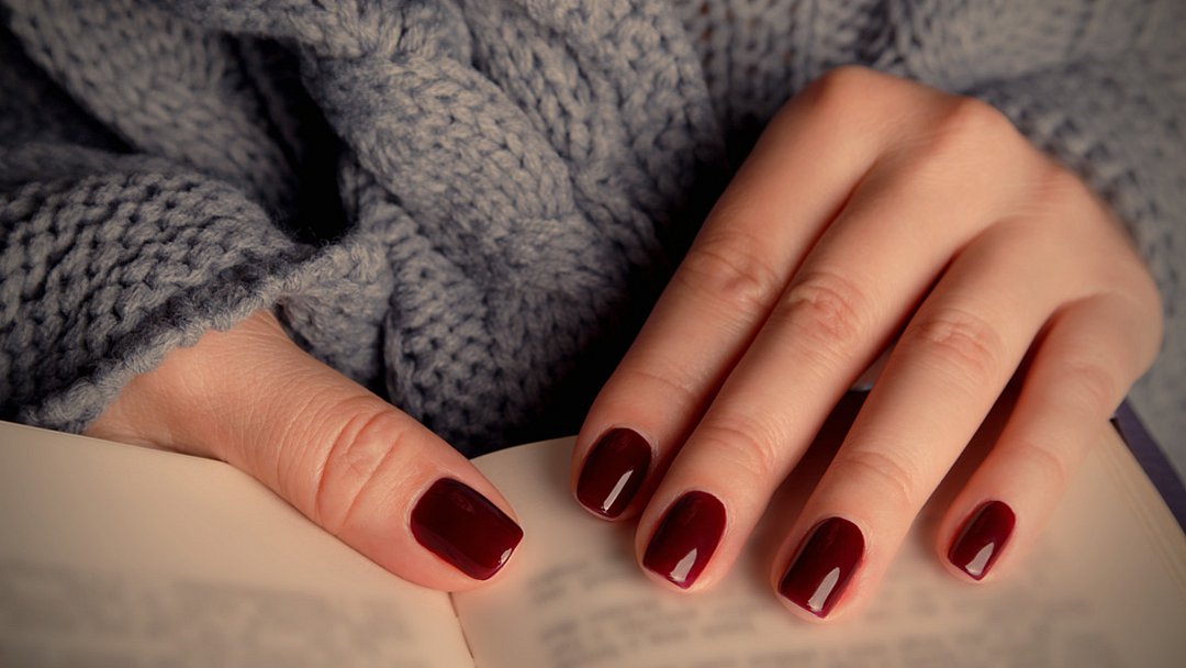 Eine Person mit dunkelroten, lackierten Fingernägeln hält ein aufgeschlagenes Buch in der Hand und trägt einen grob gestrickten grauen Pullover.