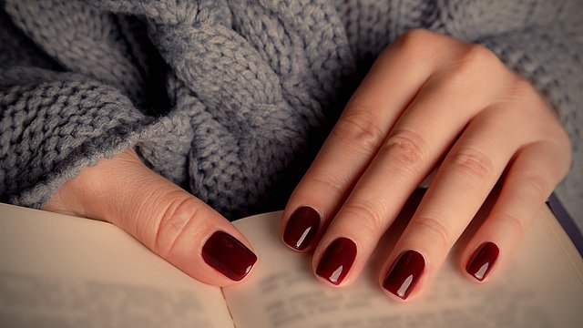 Eine Person mit dunkelroten, lackierten Fingernägeln hält ein aufgeschlagenes Buch in der Hand und trägt einen grob gestrickten grauen Pullover.