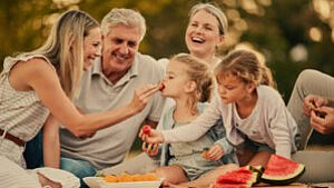 Eine fünfköpfige Familie, darunter zwei Kinder, sitzt an einem sonnigen Tag draußen auf einer Picknickdecke, isst gemeinsam Obst und lacht. Ein Erwachsener füttert ein kleines Mädchen, während ein anderes Kind ein Stück Wassermelone isst.