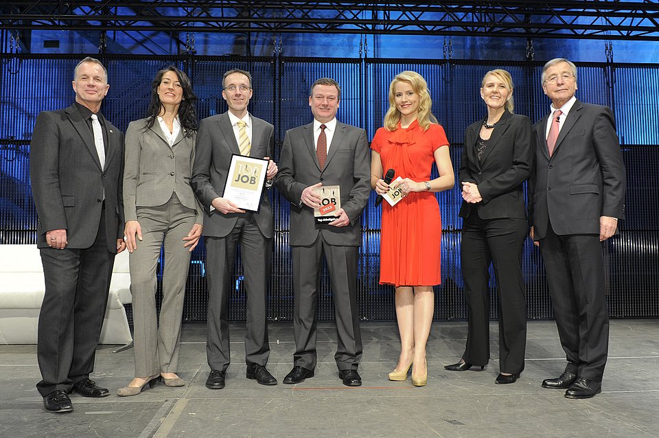 Top Job-Preisverleihung 2012 Sieben formell gekleidete Personen stehen gemeinsam auf einer Bühne, einige halten Auszeichnungen in der Hand und lächeln in die Kamera.