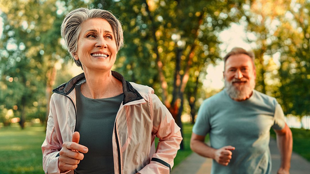 Eine lächelnde ältere Frau und ein bärtiger Mann joggen gemeinsam auf einem Parkweg, umgeben von Bäumen und Grün, in warmes Sonnenlicht getaucht.
