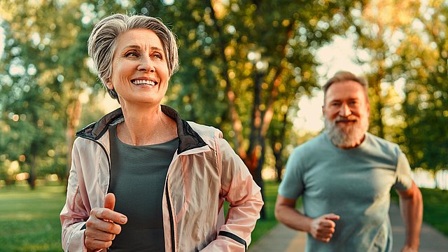 Eine lächelnde ältere Frau und ein bärtiger Mann joggen gemeinsam auf einem Parkweg, umgeben von Bäumen und Grün, in warmes Sonnenlicht getaucht.