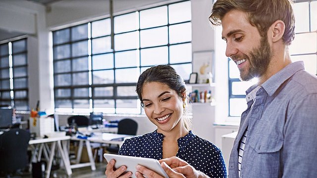 Zwei Personen stehen in einem modernen Büro und lächeln, während sie gemeinsam auf ein Tablet schauen. Im Hintergrund sind Schreibtische, Stühle und große Fenster zu sehen, durch die Tageslicht hereinfällt.