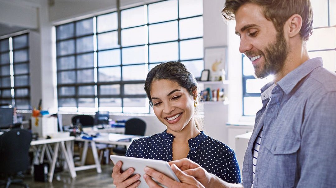 Zwei Personen stehen in einem modernen Büro und lächeln, während sie gemeinsam auf ein Tablet schauen. Im Hintergrund sind Schreibtische, Stühle und große Fenster zu sehen, durch die Tageslicht hereinfällt.