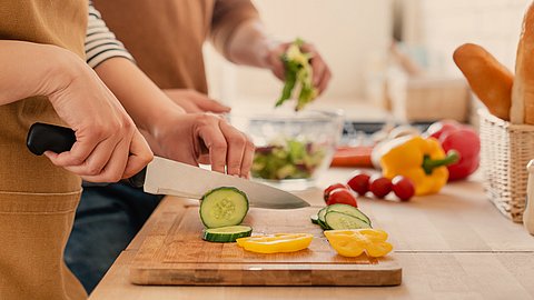 Zwei Personen bereiten gemeinsam in einer Küche Essen zu und schneiden dabei auf einem Holzschneidebrett frisches Gemüse wie Gurken und gelbe Paprika in Scheiben. Auf der Arbeitsplatte sind weiteres Gemüse und Brot zu sehen.