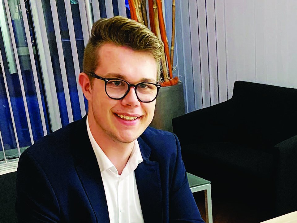 Ein junger Mann mit Brille und Anzug lächelt, während er an einem Schreibtisch in einer modernen Bürolounge sitzt.