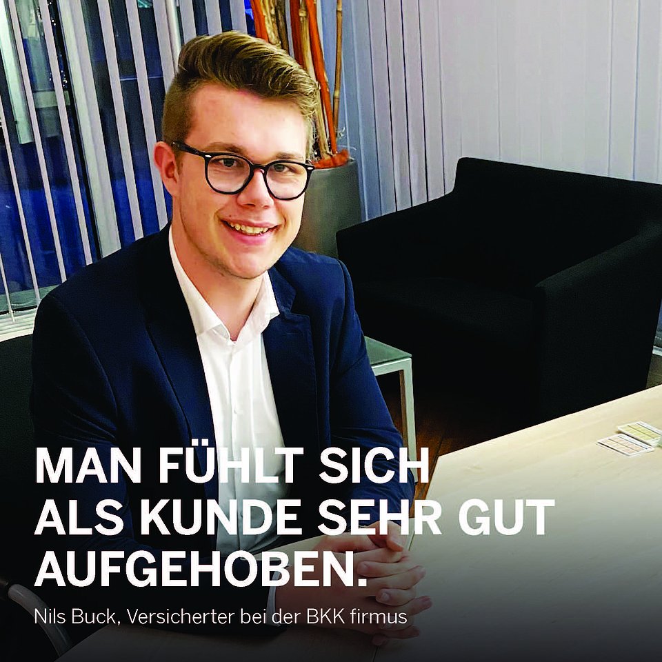 Ein junger Mann mit Brille und Anzug lächelt, während er an einem Schreibtisch in einer modernen Bürolounge sitzt.
