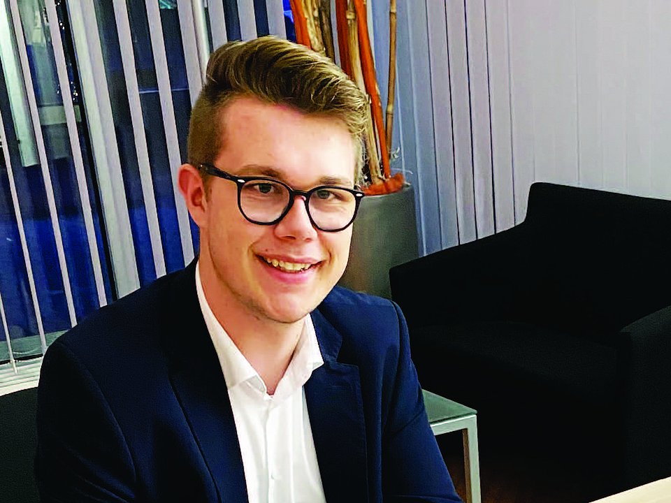 Ein junger Mann mit Brille und Anzug lächelt, während er an einem Schreibtisch in einer modernen Bürolounge sitzt.