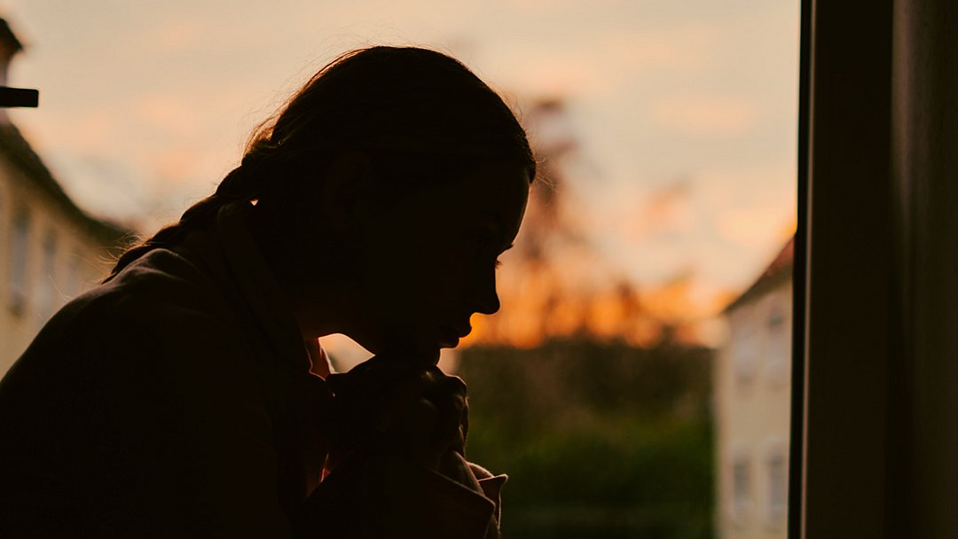 Silhouette einer Person, die drinnen sitzt, sich nach vorne beugt, die Hände unter dem Kinn verschränkt hält und nach draußen auf den Sonnenuntergang blickt. Der Himmel ist orangefarben, und im Hintergrund sind Gebäude zu sehen.