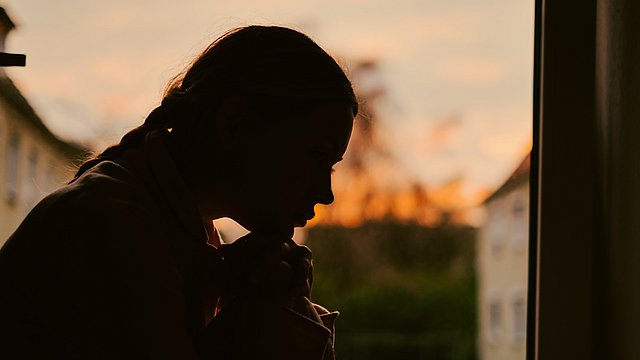 Silhouette einer Person, die drinnen sitzt, sich nach vorne beugt, die Hände unter dem Kinn verschränkt hält und nach draußen auf den Sonnenuntergang blickt. Der Himmel ist orangefarben, und im Hintergrund sind Gebäude zu sehen.