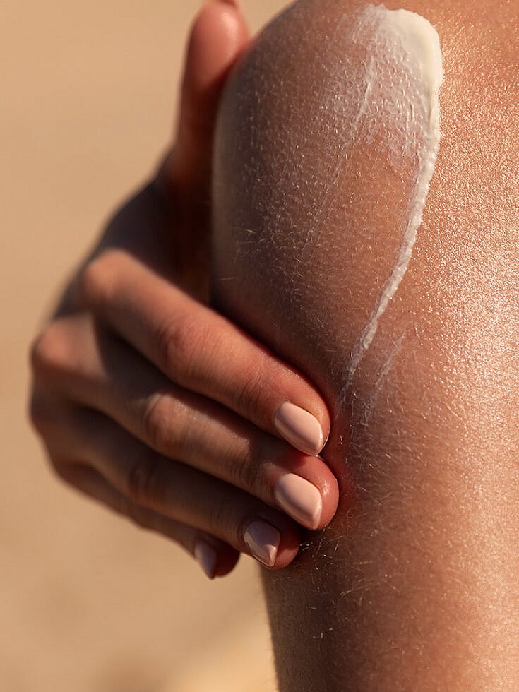 Eine Nahaufnahme einer Person, die weiße Sonnencreme auf ihre Schulter aufträgt, wobei ihre gepflegten Finger die Creme sanft auf der sonnenbeschienenen Haut verteilen.