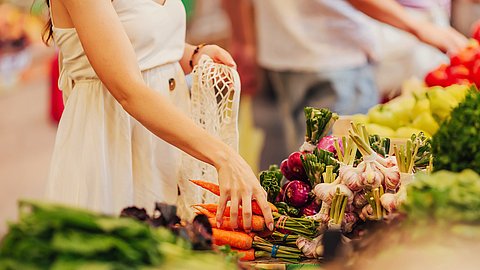 Eine Person in einem weißen Kleid greift an einem Marktstand, der mit Gemüse wie Blattgemüse, violetten Zwiebeln und Knoblauch gefüllt ist, nach frischen Möhren.