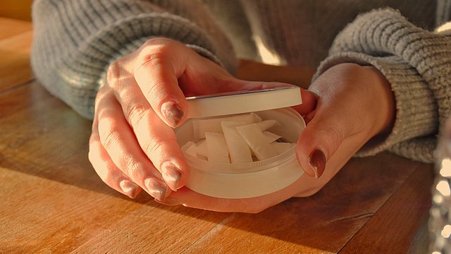 Eine Person in einem grauen Pullover hält einen offenen, runden Behälter in der Hand, in dem sich mehrere weiße Beutel befinden; der Behälter steht auf einem Holztisch im warmen Sonnenlicht.