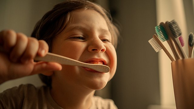 Ein kleines Kind, das lächelt und sich die Zähne putzt, hält eine hölzerne Zahnbürste in der Hand. Rechts ist ein Becher mit mehreren bunten Zahnbürsten zu sehen, und aus einem nahegelegenen Fenster fällt warmes Licht herein.