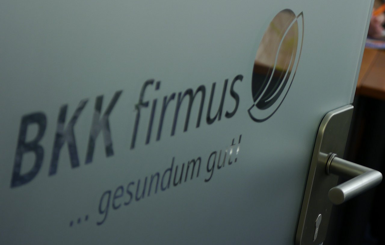 Milchglastür mit BKK firmus ...gesundum gut! und einem Logo, teilweise geöffnet mit sichtbarem Griff.