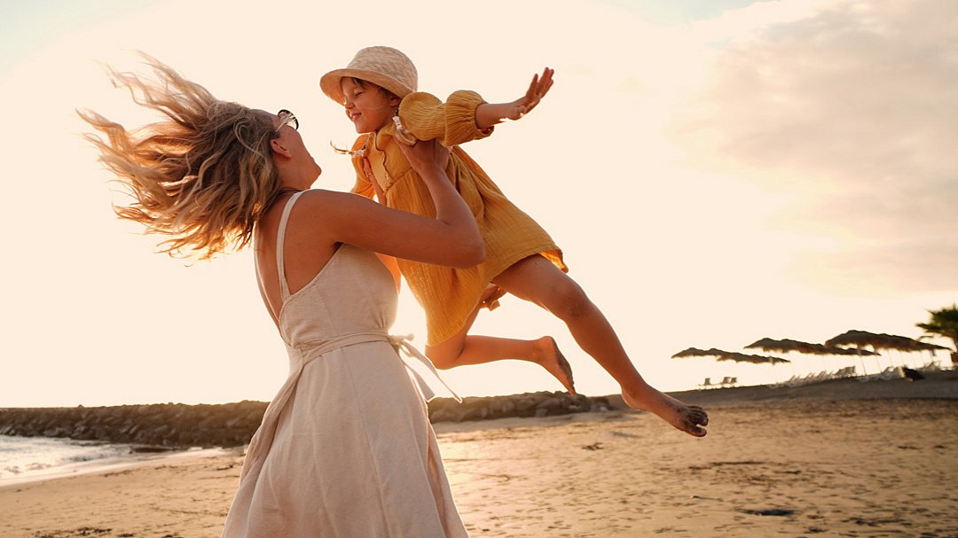 Eine Frau in einem hellen Kleid hebt ein lächelndes Kind in einem gelben Outfit und mit einem gelben Hut an einem Sandstrand bei Sonnenuntergang hoch und wirbelt es herum; beide wirken fröhlich und unbeschwert.