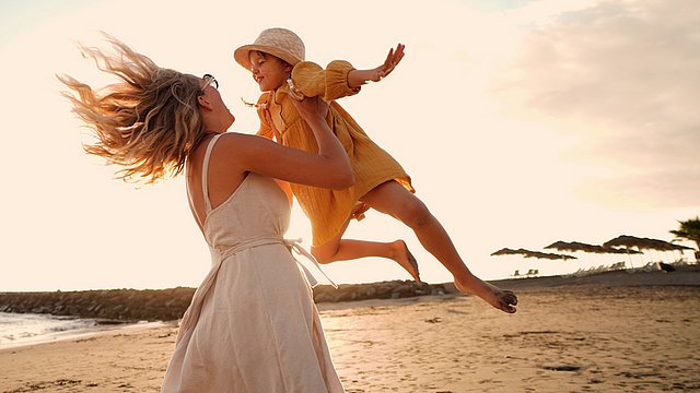 Eine Frau in einem hellen Kleid hebt ein lächelndes Kind in einem gelben Outfit und mit einem gelben Hut an einem Sandstrand bei Sonnenuntergang hoch und wirbelt es herum; beide wirken fröhlich und unbeschwert.