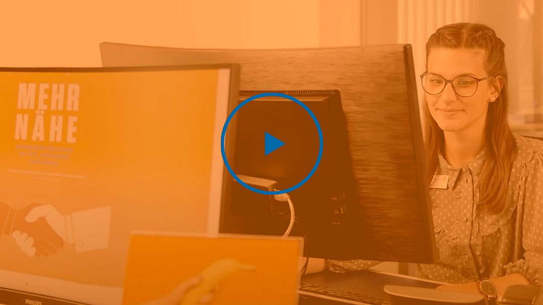 Eine Frau mit Brille sitzt an einem Schreibtisch und arbeitet am Computer. Auf dem Bildschirm vor ihr ist der Schriftzug „MEHR NÄHE“ zu sehen. Das Bild ist mit einem orangefarbenen Filter versehen, und in der Mitte ist eine Wiedergabetaste eingeblendet.