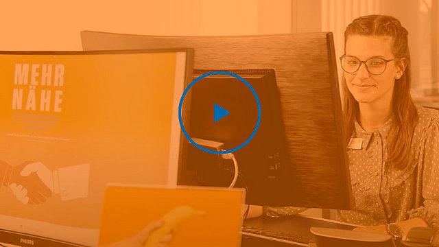 Eine Frau mit Brille sitzt an einem Schreibtisch und arbeitet am Computer. Auf dem Bildschirm vor ihr ist der Schriftzug „MEHR NÄHE“ zu sehen. Das Bild ist mit einem orangefarbenen Filter versehen, und in der Mitte ist eine Wiedergabetaste eingeblendet.