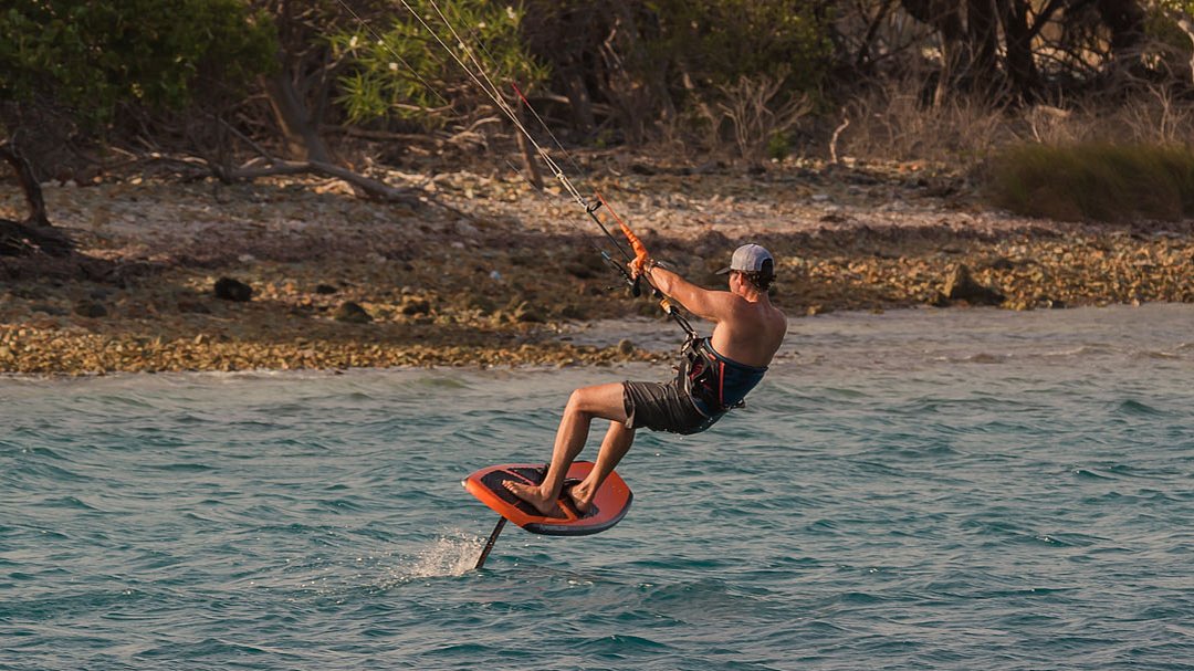 Eine Person fährt auf einem Hydrofoil-Board beim Kitesurfen in der Nähe einer felsigen Küste und trägt dabei eine Mütze, eine Sonnenbrille und eine Badehose. Im Hintergrund sind Bäume und Sträucher zu sehen.