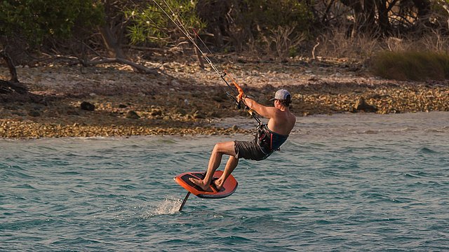 Eine Person fährt auf einem Hydrofoil-Board beim Kitesurfen in der Nähe einer felsigen Küste und trägt dabei eine Mütze, eine Sonnenbrille und eine Badehose. Im Hintergrund sind Bäume und Sträucher zu sehen.