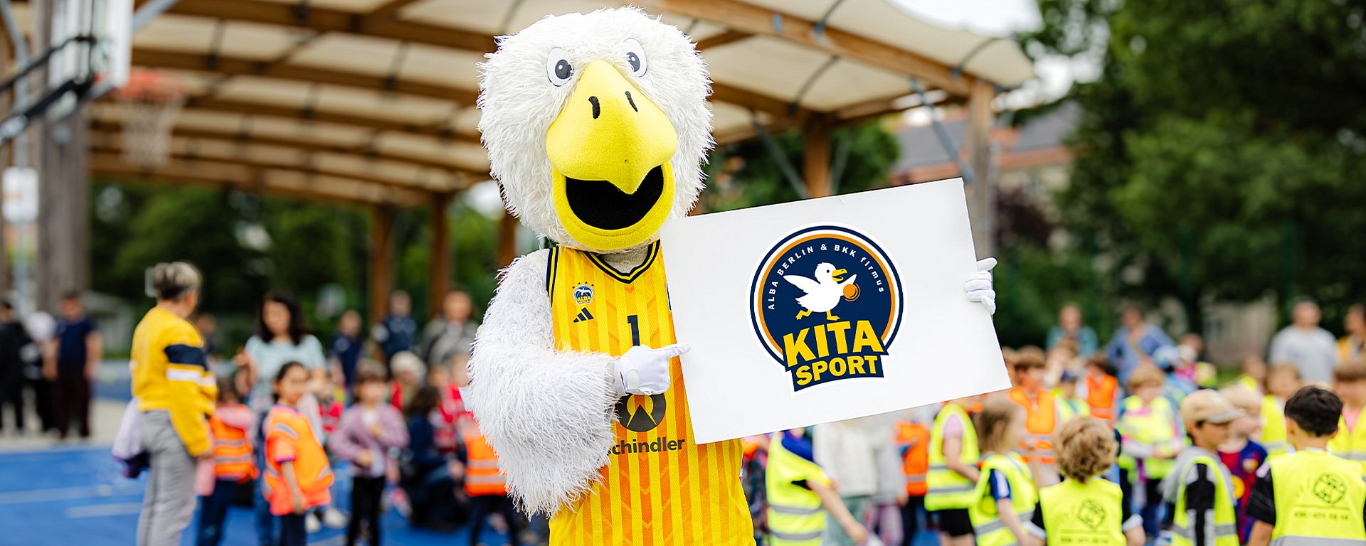 Das Maskottchen von ALBA Berlin hält bei einer Freiluftveranstaltung mit Kindern das Logo für den Kita-Sport hoch.