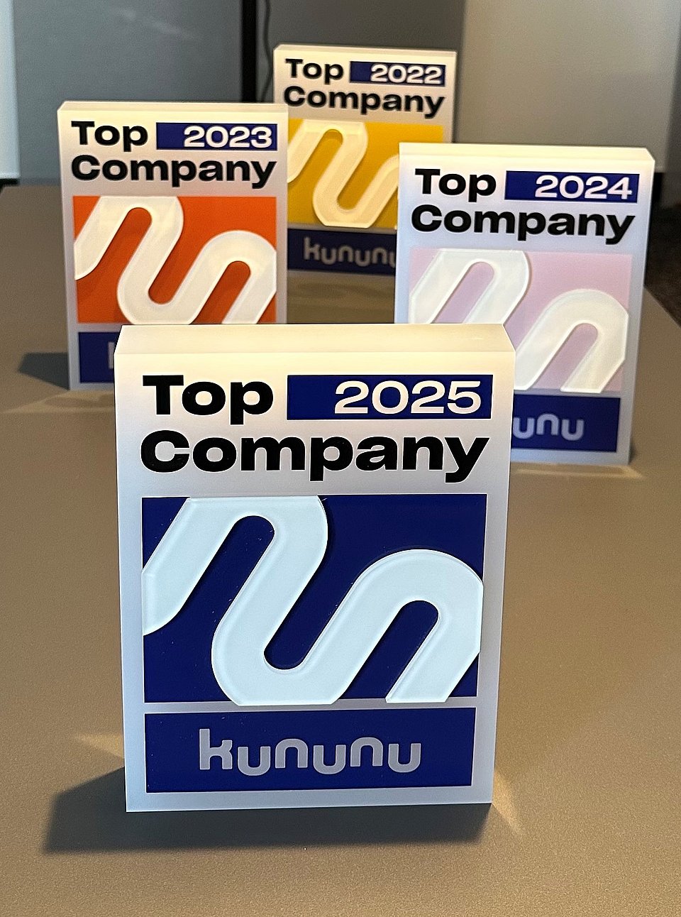 kununu Top Company Vier Top Company Awards von kununu für 2023, 2024 und 2025 auf einem Tisch.