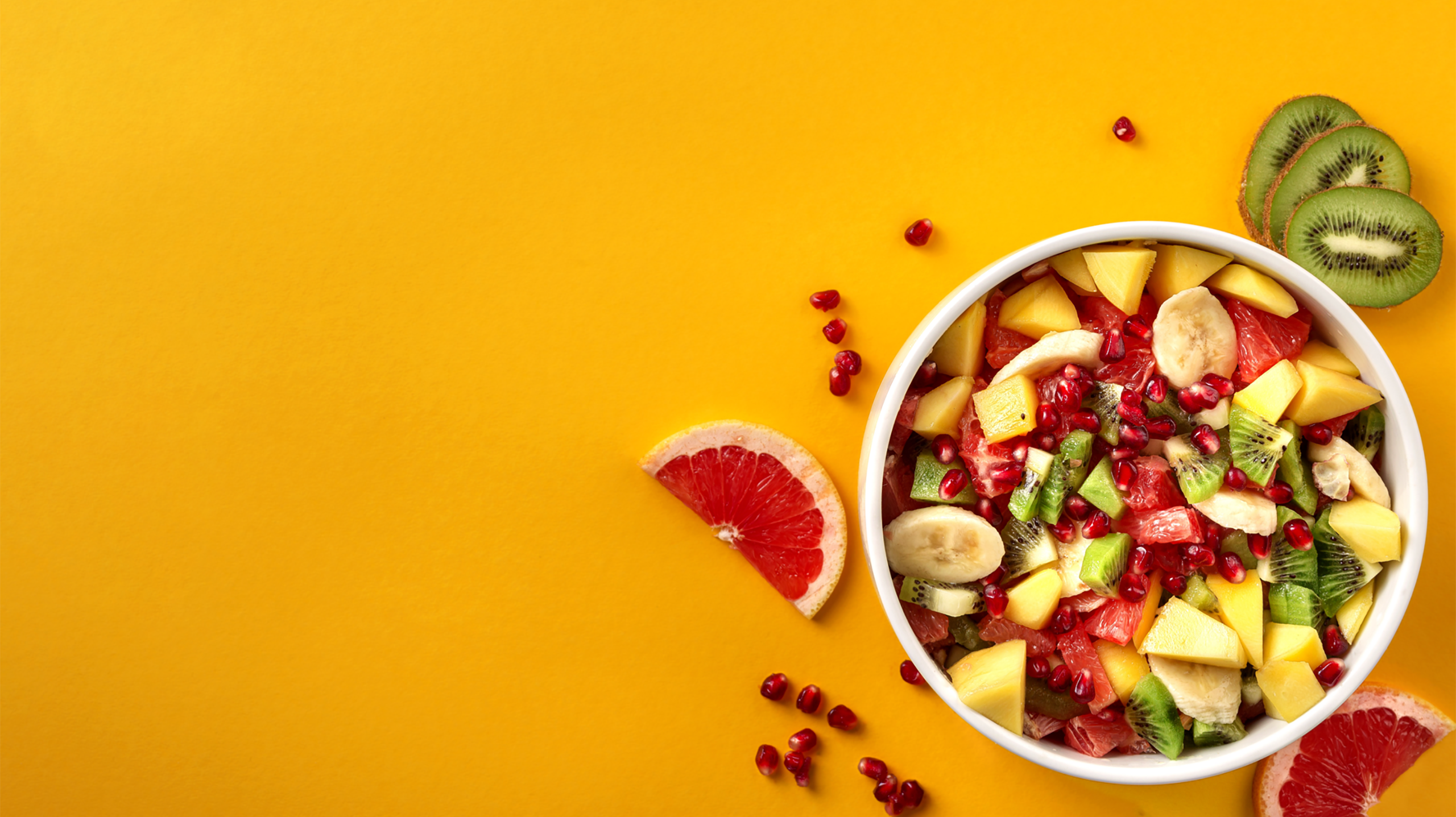 Schüssel mit gemischtem Obstsalat mit Banane, Kiwi, Mango und Granatapfel auf einem gelben Hintergrund.