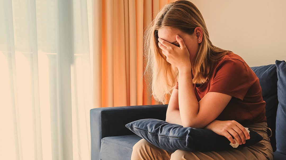 Eine Frau sitzt auf einem blauen Sofa, den Kopf in einer Hand, das Gesicht verborgen, und wirkt verzweifelt. Sie hält ein Kissen auf dem Schoß, und Sonnenlicht fällt durch die Vorhänge in der Nähe herein.