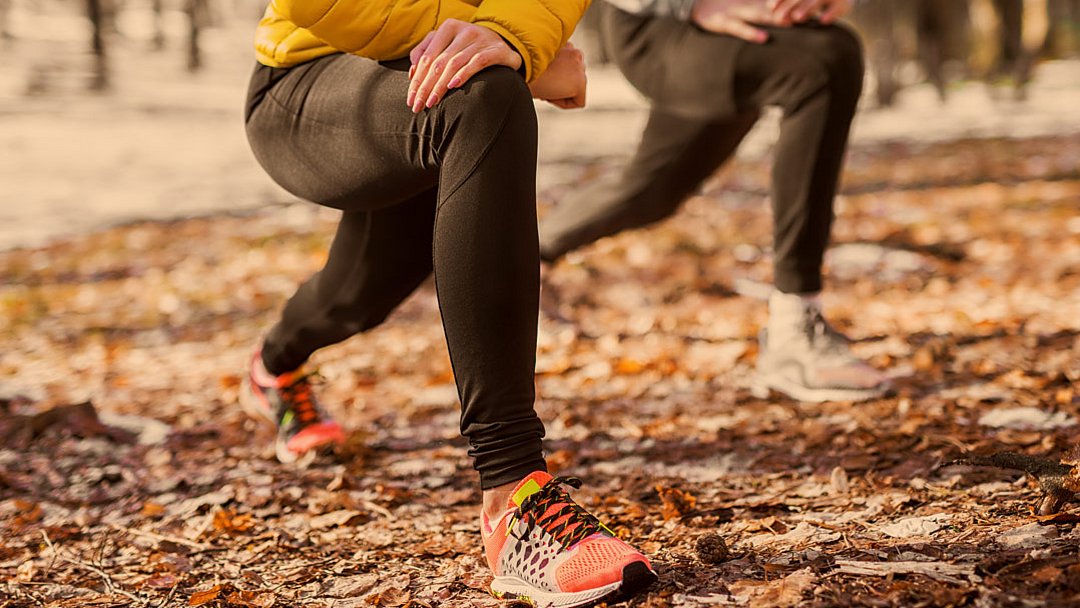 Zwei Personen in Sportkleidung und Turnschuhen machen im Freien auf einem mit Laub bedeckten Waldboden Ausfallschritte. Der Fokus liegt auf ihren Beinen und Füßen; eine der beiden Personen trägt eine gelbe Jacke und bunte Schuhe.