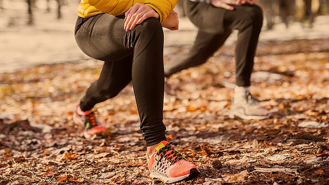Zwei Personen in Sportkleidung und Turnschuhen machen im Freien auf einem mit Laub bedeckten Waldboden Ausfallschritte. Der Fokus liegt auf ihren Beinen und Füßen; eine der beiden Personen trägt eine gelbe Jacke und bunte Schuhe.