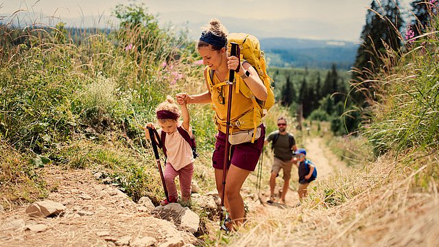 Eine Frau und ein kleines Kind wandern mit Wanderstöcken einen felsigen Pfad hinauf, gefolgt von zwei Personen im Hintergrund. Die Landschaft besteht aus Gras, Wildblumen und Bäumen in der Ferne unter einem teilweise bewölkten Himmel.