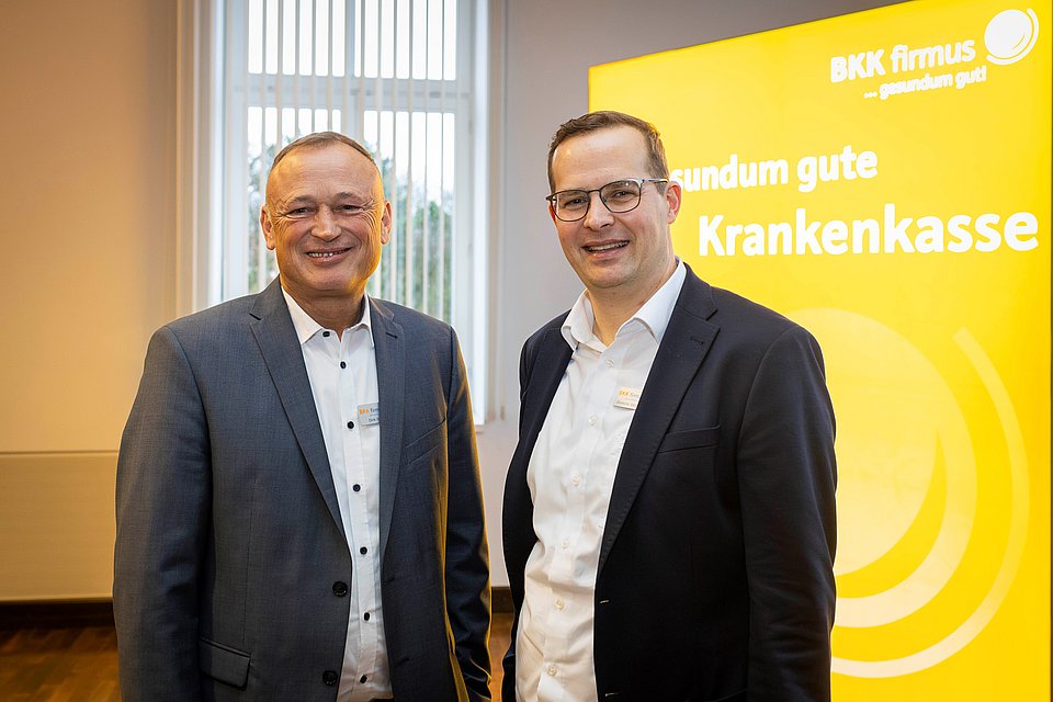 Zwei Männer in Geschäftskleidung stehen lächelnd vor einem leuchtend gelben Schild der BKK firmus Krankenkasse.