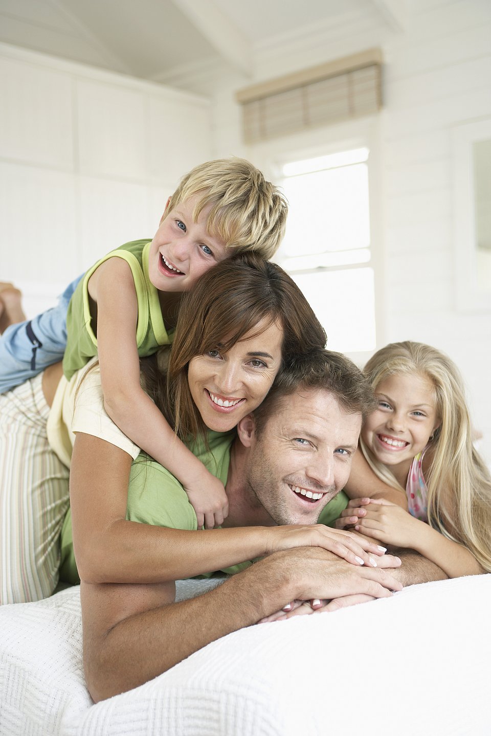Lächelnde vierköpfige Familie, die auf einem Bett liegt, wobei die Kinder ihre Eltern spielerisch umarmen.