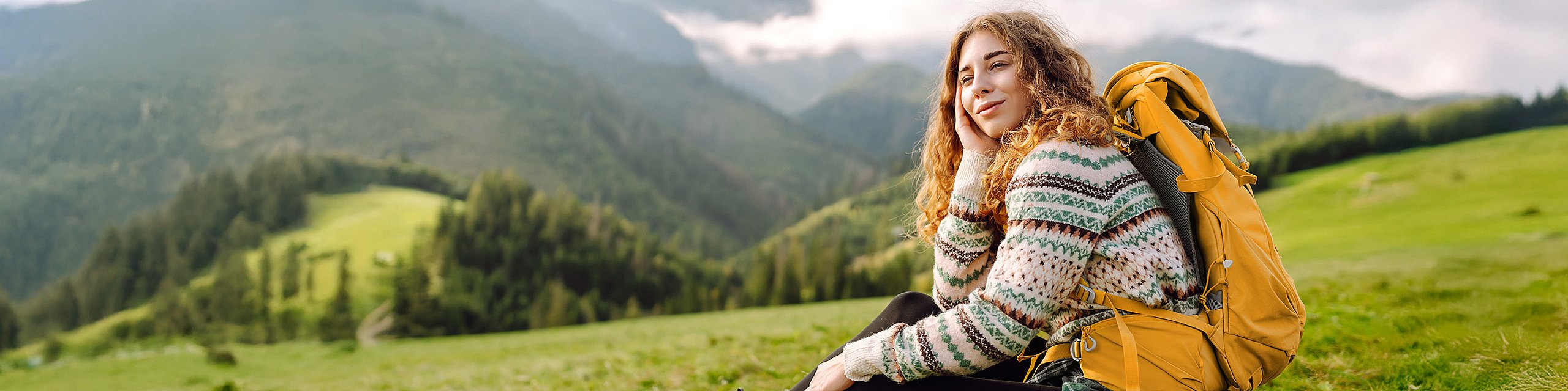 Eine junge Frau mit Rucksack und Wanderstiefeln sitzt auf einer Wiese in den Bergen, sie lächelt.
