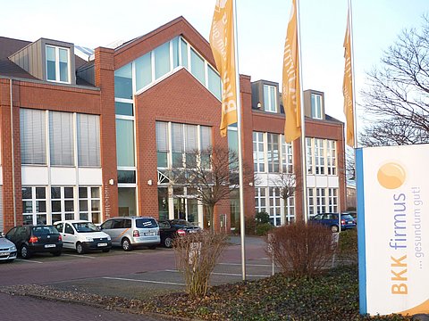Bürogebäude aus Backstein mit großen Fenstern, geparkten Autos und dem Schild und den Fahnen der BKK firmus davor.