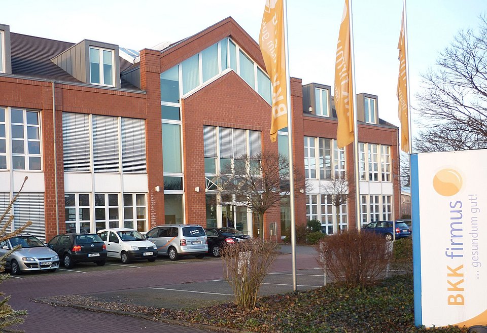 Bürogebäude aus Backstein mit großen Fenstern, geparkten Autos und dem Schild und den Fahnen der BKK firmus davor.