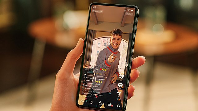 Eine Hand hält ein Smartphone, auf dessen Bildschirm ein Live-Video eines lächelnden jungen Mannes zu sehen ist, der in die Kamera zeigt; auf dem Bildschirm sind Kommentare und Reaktionen zu sehen. Im Hintergrund sind ein verschwommener Tisch und Stühle zu erkennen.