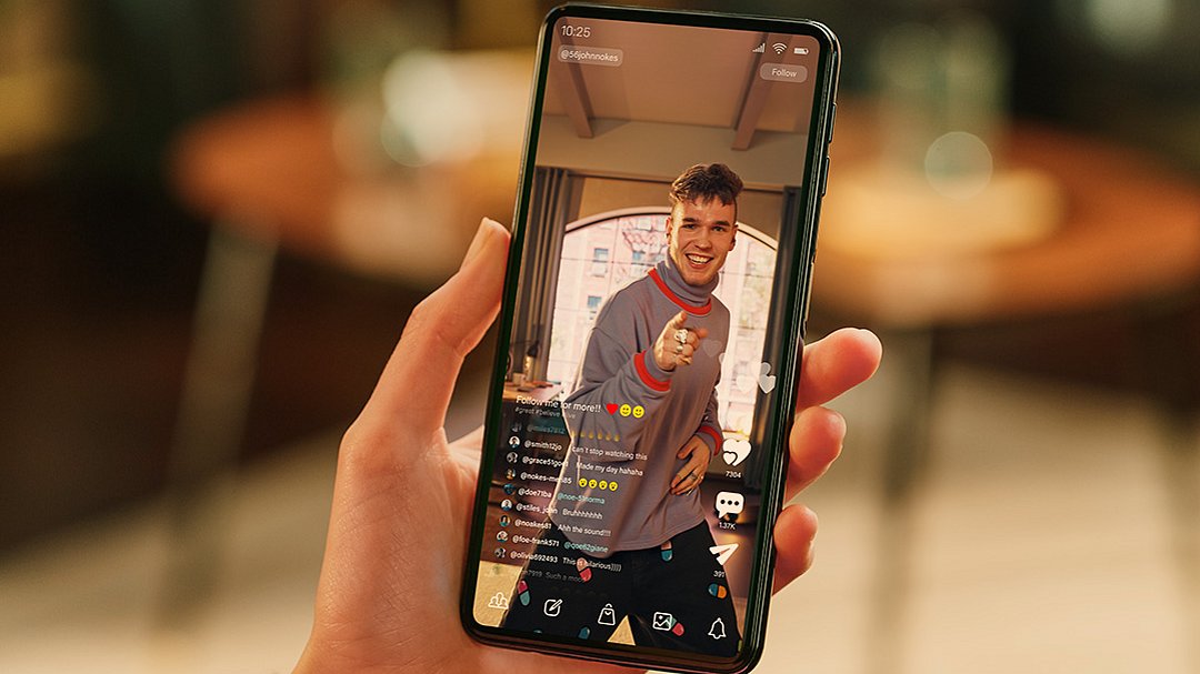 Eine Hand hält ein Smartphone, auf dessen Bildschirm ein Live-Video eines lächelnden jungen Mannes zu sehen ist, der in die Kamera zeigt; auf dem Bildschirm sind Kommentare und Reaktionen zu sehen. Im Hintergrund sind ein verschwommener Tisch und Stühle zu erkennen.