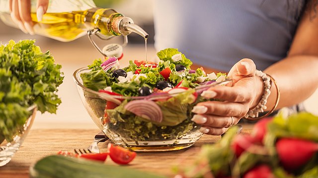 Eine Person gießt Olivenöl über einen frischen Salat in einer Glasschüssel. Der Salat besteht aus Kopfsalat, Tomaten, roten Zwiebeln, Gurken, Oliven und Feta-Käse. Auf dem Tisch stehen weitere Gemüsesorten.