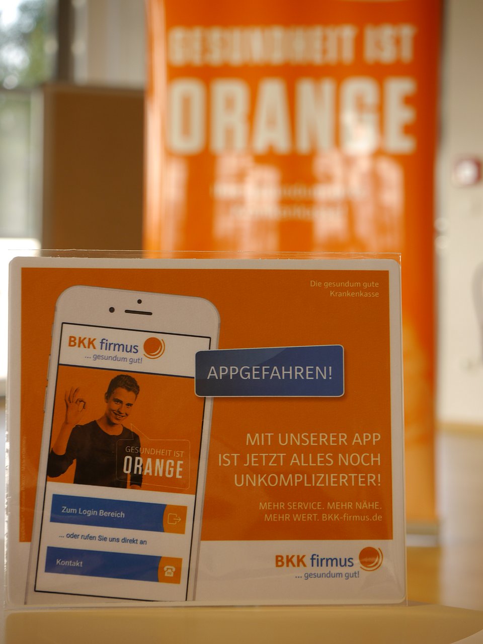 BKK firmus App Poster und Flyer für eine Gesundheits-App, mit orangefarbenem und weißem Text und einem Smartphone-Bild.
