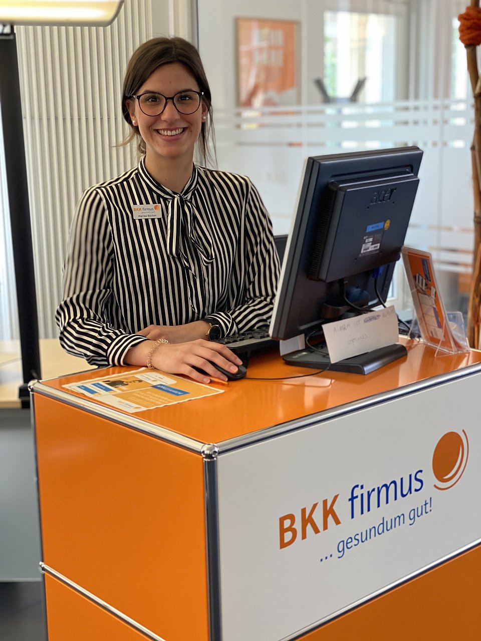 Eine Frau mit Brille lächelt an einem Empfangstresen mit Computer in der BKK firmus-Geschäftsstelle.