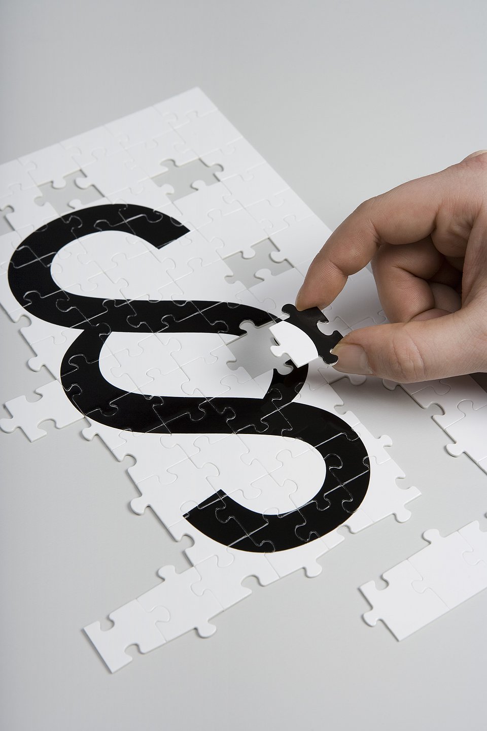 Eine Hand setzt ein Puzzleteil in ein Puzzle ein, das ein schwarzes Absatzsymbol (§) auf weißem Grund zeigt.