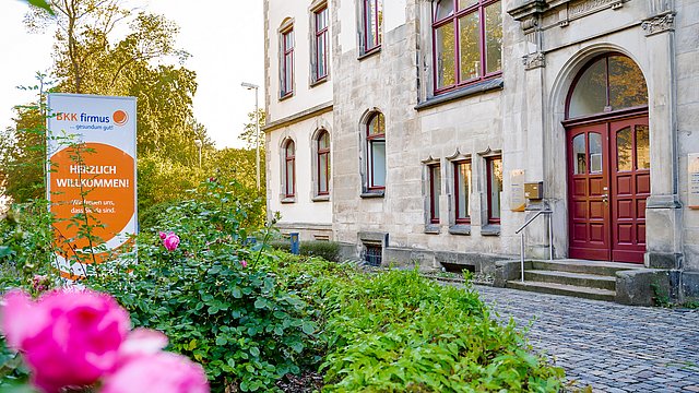 Ein historisches Steinhaus mit hohen Fenstern und einer roten Tür, umgeben von blühenden rosa Rosen. Neben dem Weg, der zum Eingang führt, steht ein Schild auf Deutsch mit der Aufschrift „Herzlich Willkommen!“.