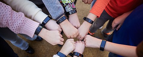 Eine Gruppe von Kindern steht im Freien im Kreis, jeder streckt seine Faust in Richtung Mitte aus, und alle tragen Armbänder am Handgelenk.