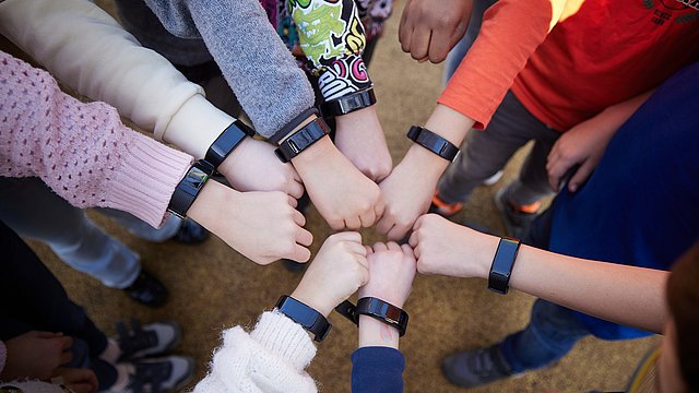 Eine Gruppe von Kindern steht im Freien im Kreis, jeder streckt seine Faust in Richtung Mitte aus, und alle tragen Armbänder am Handgelenk.