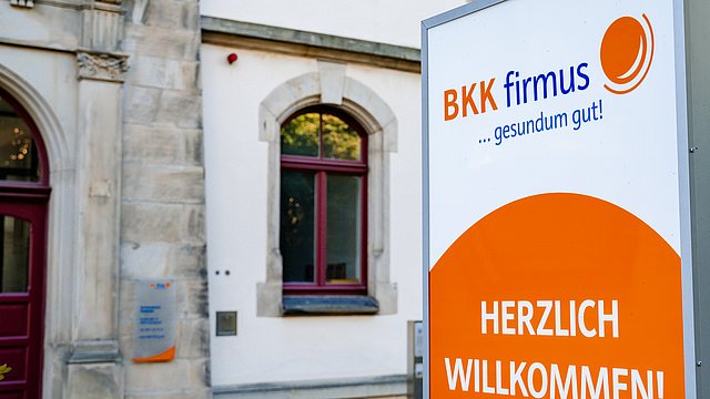 Ein Schild der Krankenkasse „BKK firmus“ heißt Besucher vor einem Gebäude mit Bogenfenstern willkommen.