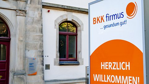 Ein Schild der Krankenkasse „BKK firmus“ heißt Besucher vor einem Gebäude mit Bogenfenstern willkommen.