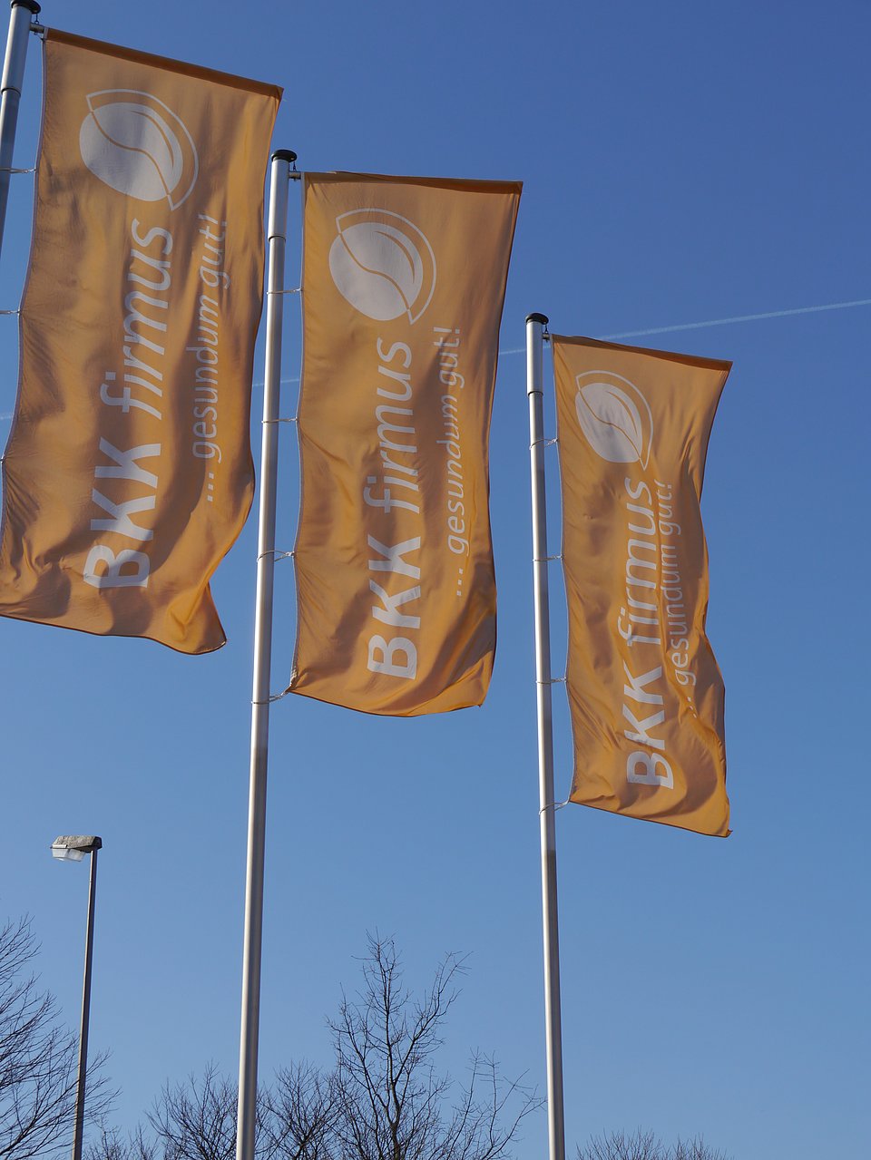 Drei orangefarbene BKK firmus-Flaggen wehen an Masten vor einem strahlend blauen Himmel.