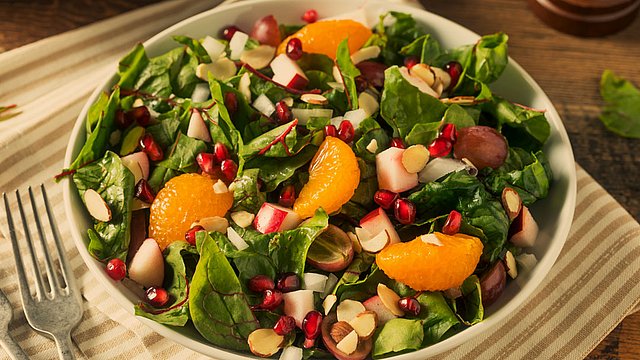 Eine Schüssel mit frischem Salat aus Spinat, Mandarinenstücken, Granatapfelkernen, Radieschen, Weintrauben und Mandelscheiben auf einem Holztisch, daneben eine gestreifte Serviette und zwei Gabeln.